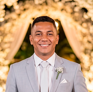 Retrato em close-up do padrinho Bruno Costa, irmão do noivo, sorrindo muito em frente a um arco de flores iluminado. Ele veste nosso elegante terno cinza claro com gravata branca. Mais um cliente satisfeito com o aluguel de trajes da Ternos Curitiba.