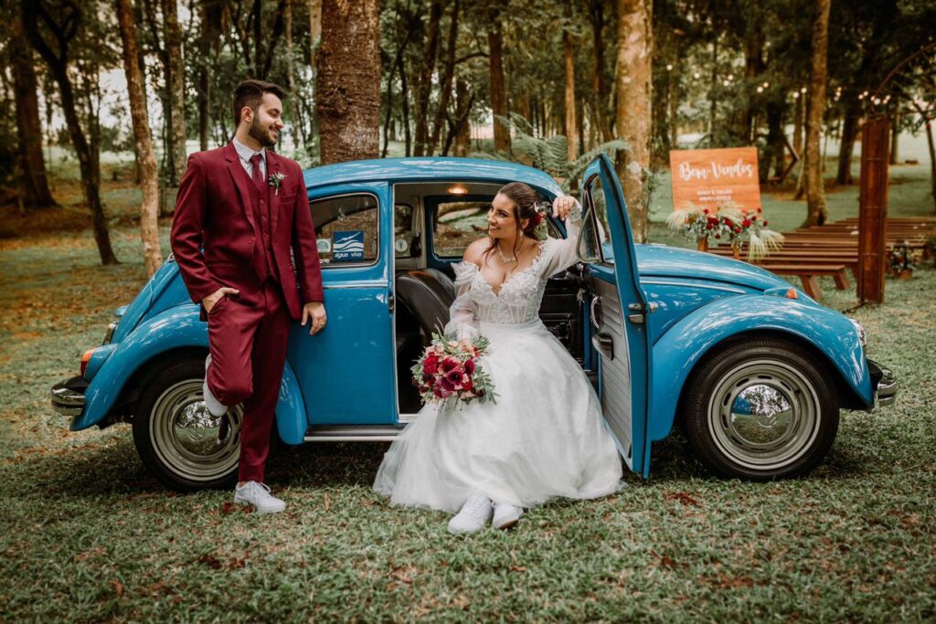 Casal de noivos sorrindo ao lado de um Fusca azul vintage em um casamento rústico ao ar livre. O noivo destaca-se vestindo um moderno terno marsala completo com colete, combinado de forma despojada com tênis branco. A melhor escolha em aluguel de terno para noivo em Curitiba.