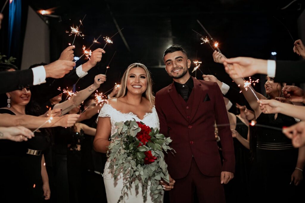 Noivos sorridentes celebrando a saída do casamento sob um corredor de faíscas (sparklers). O noivo destaca-se usando um moderno terno marsala completo, contrastando com camisa e gravata pretas. Soluções exclusivas em aluguel de terno para noivo em Curitiba.
