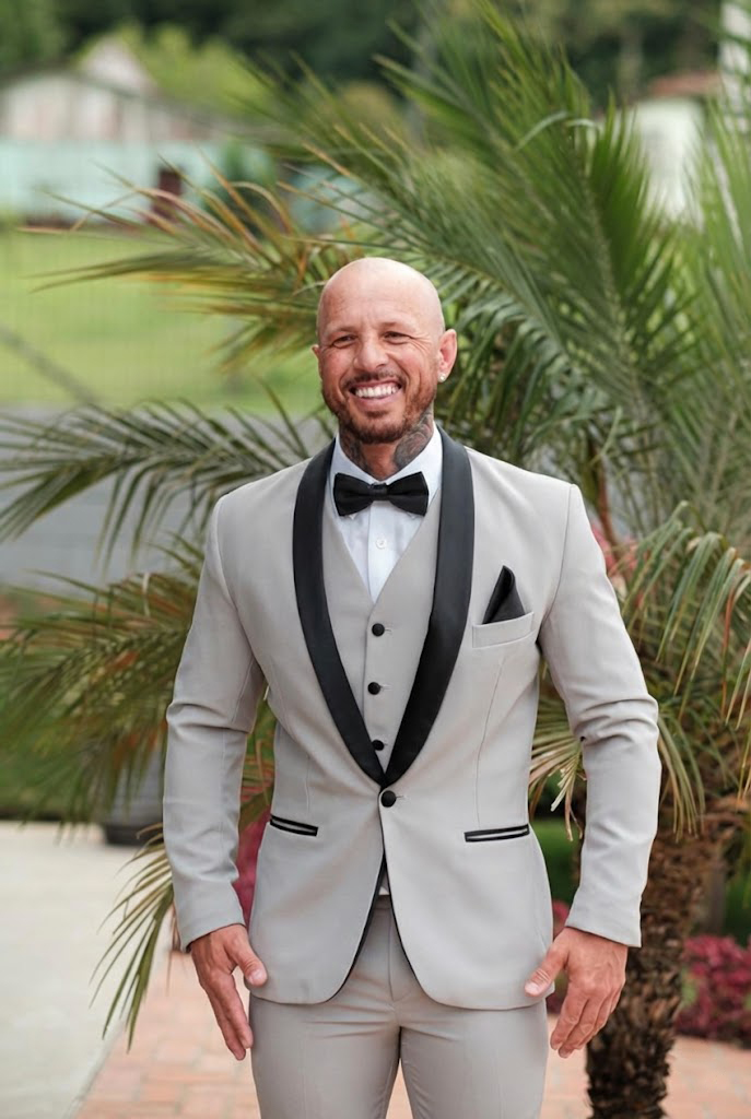 Homem sorridente ao ar livre vestindo um moderno smoking cinza claro com colete, contrastando com a lapela xale, gravata borboleta e lenço na cor preta. Excelente opção de aluguel de smoking em Curitiba para noivos ousados e eventos sofisticados.