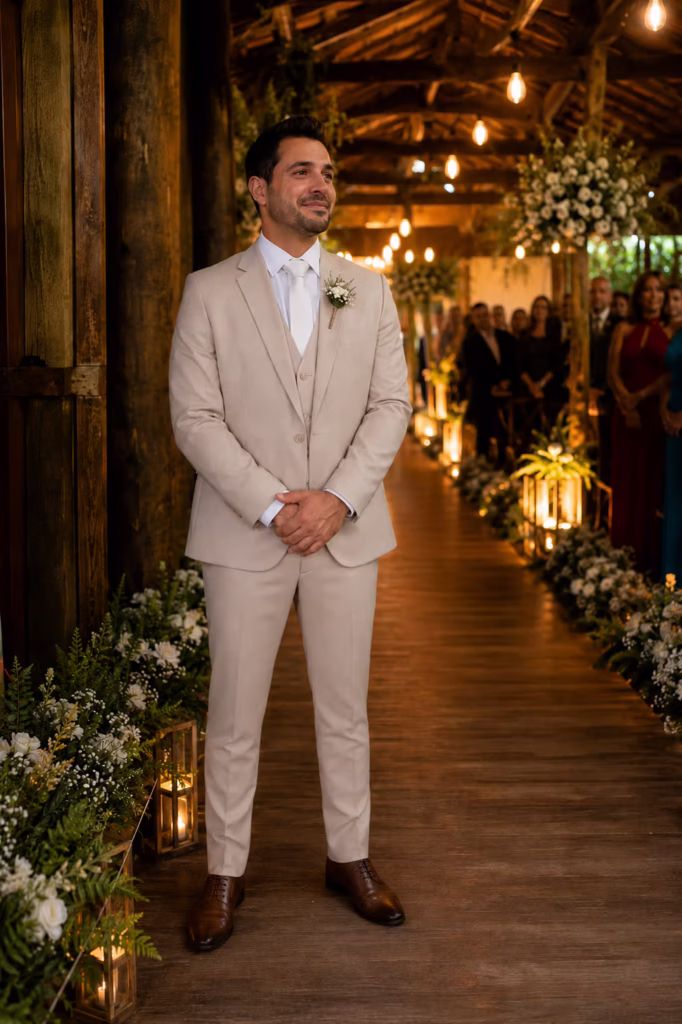 Noivo emocionado e sorridente aguardando no altar de um casamento rústico decorado com flores. Ele veste um terno cor areia de corte slim com colete, gravata branca e sapatos marrons. A escolha perfeita de aluguel de trajes para noivos em Curitiba.